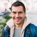 A man with short dark hair and light stubble smiles at the camera. He is wearing a blue denim jacket with a beige shearling collar over a gray sweater. In the background, there are indistinct outdoor elements and some greenery, adding a touch of freshness like after regular cleaning.