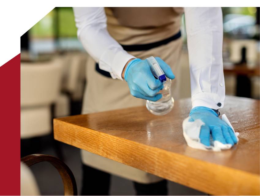 A person wearing a white shirt, black apron, and blue gloves is diligently performing healthcare cleaning by sanitizing a wooden table with a spray bottle and a white cloth. The background shows the blurred interior of what appears to be a restaurant or cafe.