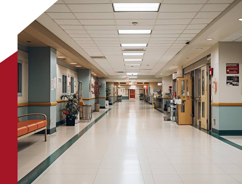 A long hospital corridor with tiled flooring, illuminated by overhead fluorescent lights. The walls are painted in light colors with a few orange accents. There is an orange bench on the left side and medical equipment stationed throughout the hallway, reflecting a commitment to meticulous healthcare cleaning standards.