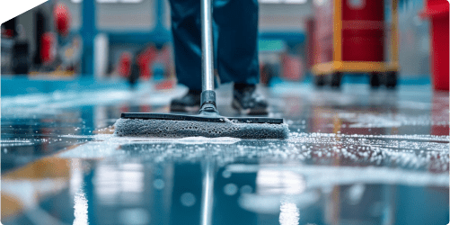 A person in dark blue pants uses a squeegee to clean a shiny, wet floor. Soap suds are visible on the surface. The background shows a large indoor manufacturing space with blurred red and yellow objects.