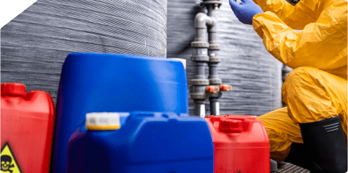 A person wearing a yellow hazmat suit and blue gloves is cleaning near several large chemical containers. Nearby, there are red and blue plastic jerrycans, one of which has a hazard symbol.