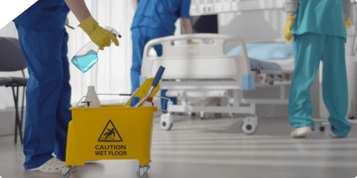 A hospital room is being cleaned. A mop bucket with a "Caution Wet Floor" sign and various cleaning supplies is in the foreground. Two healthcare professionals wearing scrubs and gloves are seen performing cleaning duties around a hospital bed in the background.