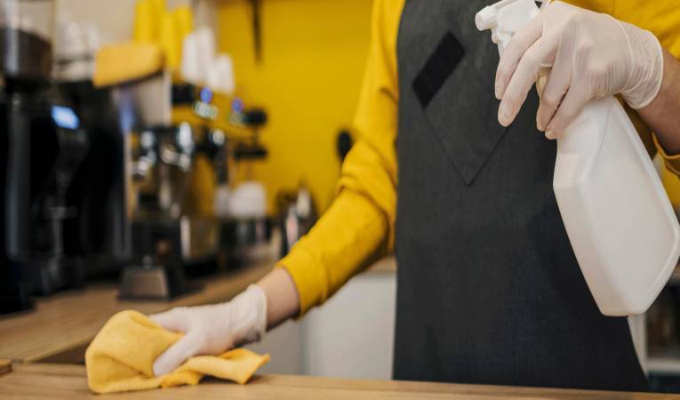 A person wearing gloves and a dark apron is meticulously cleaning a countertop, embodying top-notch restaurant sanitation standards. They hold a white spray bottle in one hand and wipe the surface with a yellow cloth. The background showcases various coffee shop equipment and vibrant yellow walls.