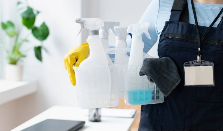 A person wearing a blue uniform and a name badge holds a plastic caddy filled with cleaning supplies, including spray bottles and a blue cleaning solution. Emphasizing cleanliness in healthcare, the individual also wears a yellow glove and holds a dark cleaning cloth. A plant is visible in the background.