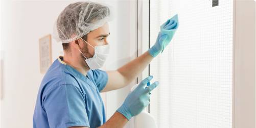 A person wearing blue scrubs, a hair net, a face mask, and gloves is diligently cleaning a glass door with a spray bottle and a blue cloth, embodying the meticulous standards of healthcare.