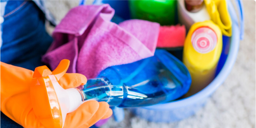 A person wearing orange gloves holds a blue spray bottle. Other cleaning supplies, including a pink cloth, a green sponge, and various bottles of cleaner, are neatly arranged inside a blue bucket. It's as if preparing for a thorough cleaning session in a kindergarten classroom.