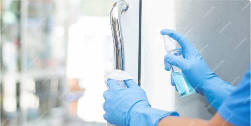 A person wearing blue gloves is cleaning a door handle with a cloth and spraying it with a cleaning solution, emphasizing the importance of hygiene in healthcare. The surroundings are blurry and out of focus.