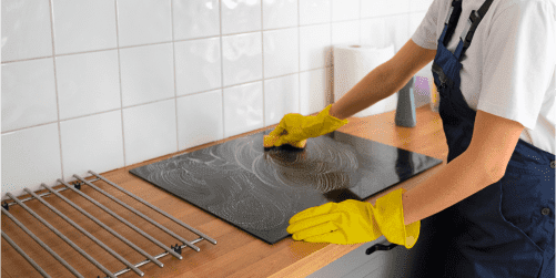 A person wearing yellow cleaning gloves is meticulously cleaning a black stove top on a wooden counter in a kitchen. Dressed in blue overalls and a white shirt, they work efficiently against the backdrop of white tiled backsplash. Nearby, a roll of paper towels stands ready for additional cleaning tasks.