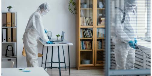 Two people in full-body protective suits and gloves are in an office-like room. One is standing at a desk with a laptop, while the other is near a window. A wooden bookshelf with various items and books is in the background, emphasizing a clean and organized healthcare environment.