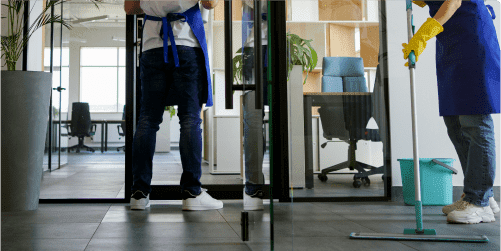 Two people in blue aprons and yellow gloves are cleaning the glass door of an office. One is mopping the floor while the other wipes down the glass. A green bucket and a chair are visible in the background, reminiscent of a kindergarten setting with large windows and plants completing the scene.