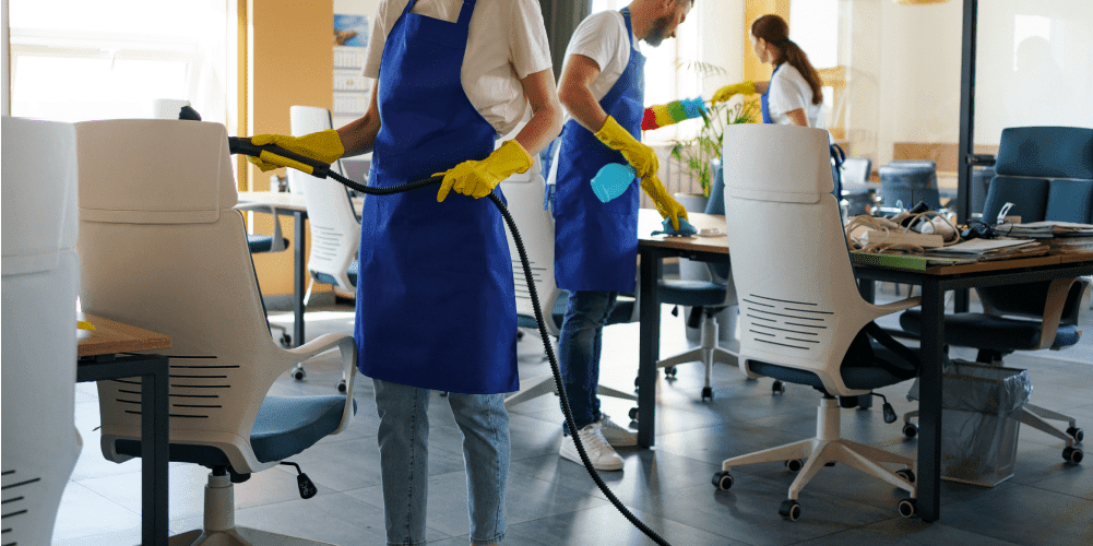 Three people are cleaning an office space, demonstrating school hygiene standards. They are wearing blue aprons and yellow gloves. One person is using a vacuum, another is using a spray bottle, and the third is wiping surfaces. The office has white chairs, desks, and a plant in the background.