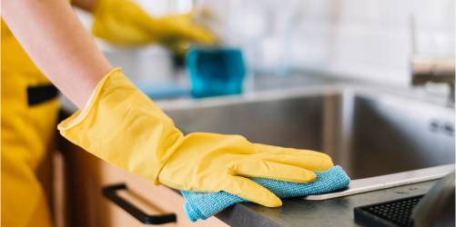 A person wearing yellow rubber gloves is wiping a kitchen counter with a blue cloth, showcasing the importance of restaurant hygiene. There is a stainless steel sink in the background along with a blurred green object, possibly a cleaning spray bottle.