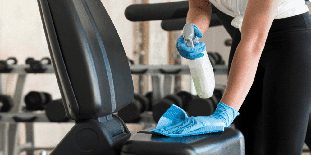 A person wearing blue gloves is cleaning exercise equipment with a spray bottle and cloth in a gym. The thorough cleaning resembles the meticulous standards of school janitorial services, with various weights and gym machines visible in the background.