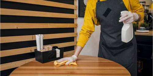 A person wearing a yellow sweater and gray apron is engaged in restaurant cleaning, diligently wiping down a round wooden table with a yellow cloth while holding a spray bottle. Nearby, there is a black container holding stir sticks, napkins, and other small items.