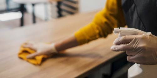 A person wearing gloves uses a spray bottle and cloth to clean a wooden surface, typical of restaurant cleaning. The individual is dressed in a yellow shirt and is partially visible from the shoulders down. The background is slightly blurred, indicating an indoor setting.