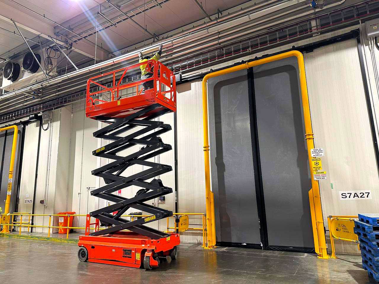 A worker stands on a raised red scissor lift, wearing a neon yellow safety vest and helmet, in an industrial warehouse. Engaged in at-height cleaning, the lift is positioned in front of a large door with yellow framing. Pipes and ducts are visible on the ceiling.