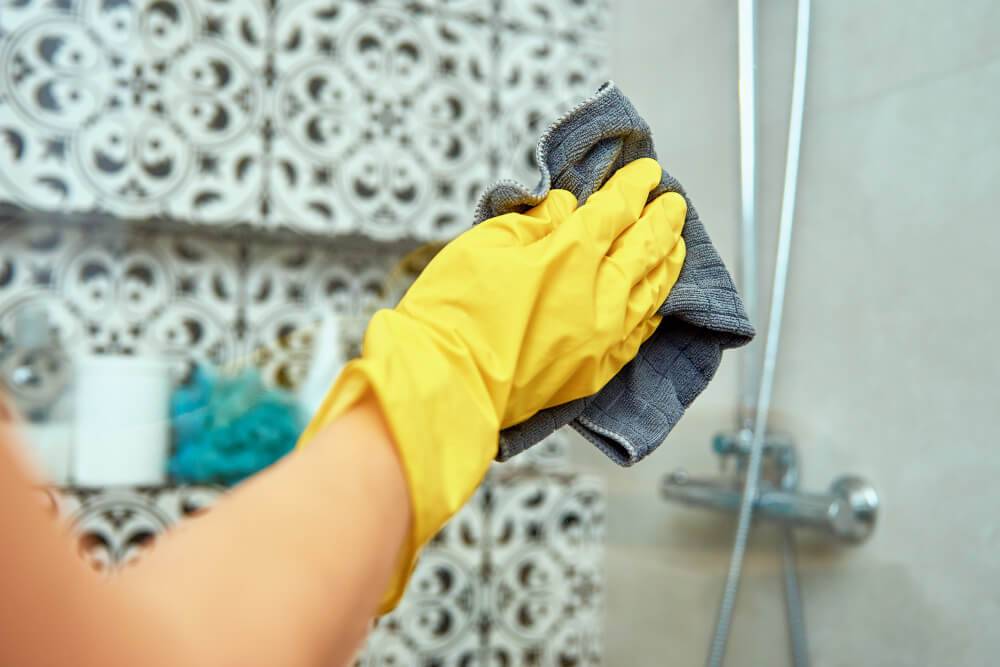 A person wearing yellow rubber gloves is engaged in regular house cleaning, using a dark cloth to clean a bathroom wall tile with a decorative pattern. A showerhead and part of a shower area are visible in the background.