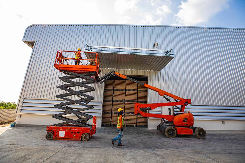 Two construction workers are in front of a large, corrugated metal building. One stands on an orange scissor lift, engaged in what appears to be at-height cleaning or inspection. The other worker remains on the ground, watching attentively.