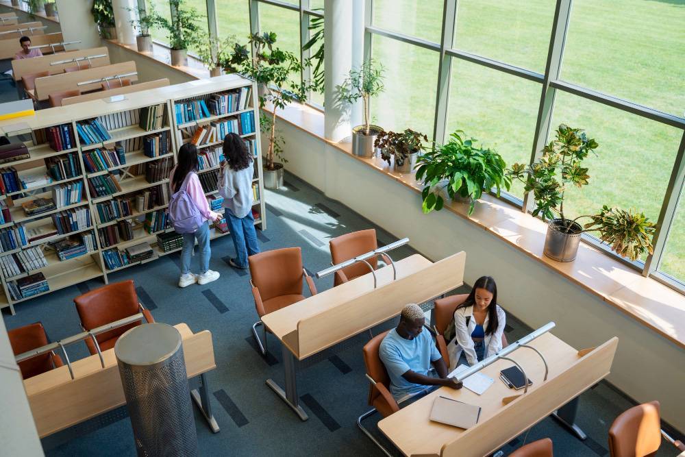 A spacious library with large windows is filled with natural light. Two people are seated at a desk with laptops, while two others stand by a bookshelf looking at books. The room, benefiting from school janitorial services, is well-lit and decorated with potted plants.