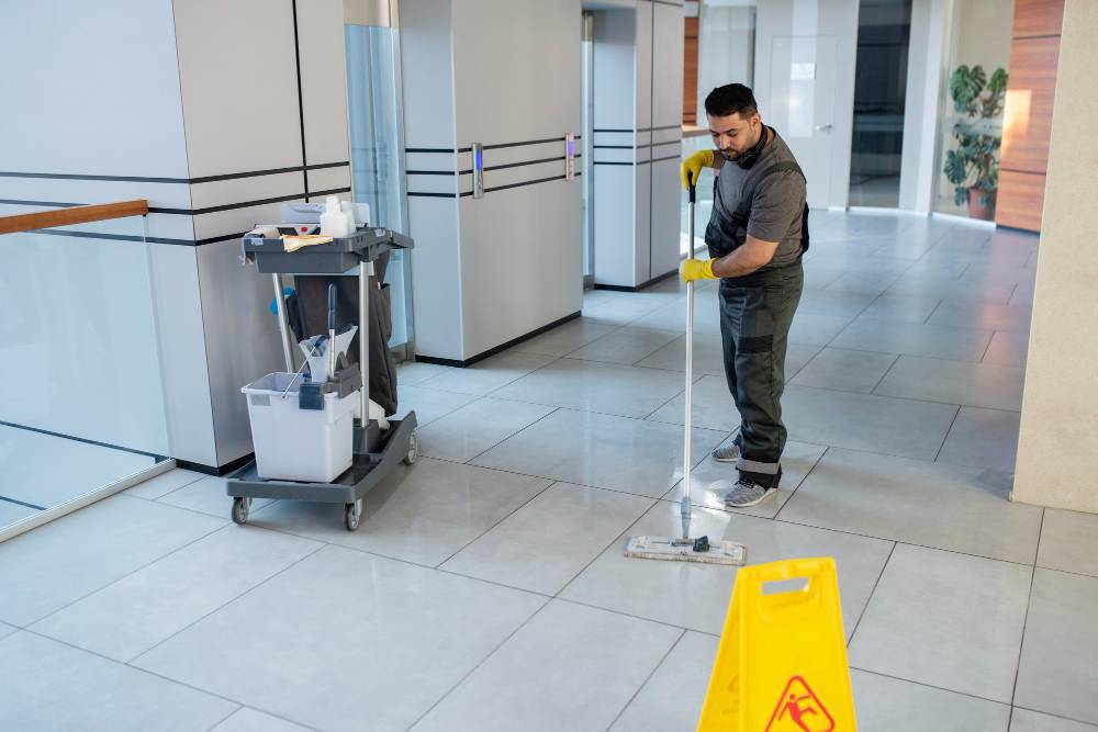 A man in gray uniform and yellow gloves is mopping the tile floor in a spacious school hallway. A cleaning cart with supplies is nearby, and a yellow caution sign indicates the wet surface. The well-lit corridor is clean and orderly.