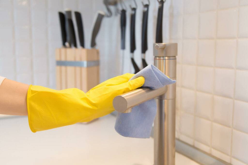 A person wearing a yellow rubber glove is cleaning a stainless steel kitchen faucet with a blue cloth, ensuring everything is spotless for the next tenant. Knives in a wooden knife block are seen in the background on a white tiled wall, reflecting the thorough move-out cleaning completed by the service.