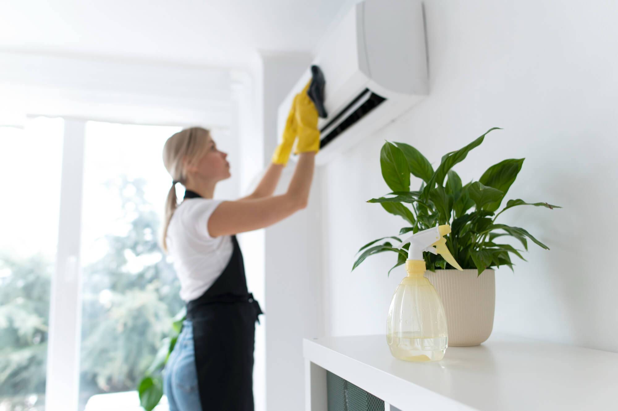 A person wearing yellow gloves and an apron cleans an air conditioning unit mounted on a wall, possibly as part of a Move-In/Move-Out Cleaning service. In the foreground, there is a white cabinet with a spray bottle and a potted plant. Sunlight streams into the room through large windows.