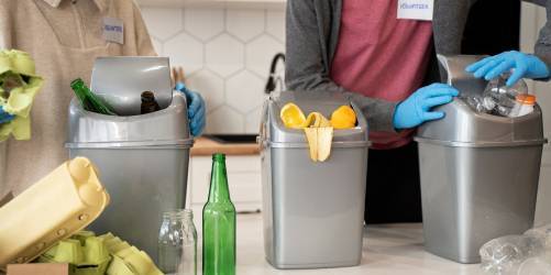 Three people at a restaurant are sorting recyclables into three grey bins labeled "glass," "plastic," and "compost." Wearing blue gloves and nametags, they carefully place bottles, cans, and biodegradable waste into the appropriate bins as part of their cleaning routine.