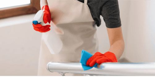 A person wearing a grey shirt, beige apron, and red gloves is cleaning a surface with a spray bottle and wiping it with a blue cloth. The scene appears to be indoors near a window, possibly in a kindergarten.