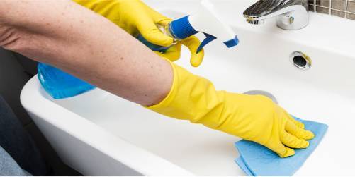 A person wearing yellow rubber gloves is cleaning a white sink with a blue cloth and a spray bottle. The hand on the left holds the spray bottle, while the hand on the right uses the cloth. This scene reflects healthcare cleaning standards, ensuring every surface meets strict hygiene criteria.