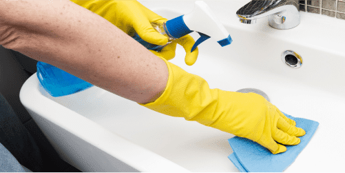 A person wearing yellow rubber gloves is cleaning a white sink, much like in a kindergarten setting. They are holding a blue spray bottle with a white nozzle in one hand and wiping the sink with a blue cloth in the other. A chrome faucet gleams in the background.