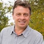 A man with short, wavy brown hair is smiling at the camera. He is wearing a light gray button-up shirt and is standing outdoors with green trees in the background, reflecting his commitment to providing top-notch school janitorial services.