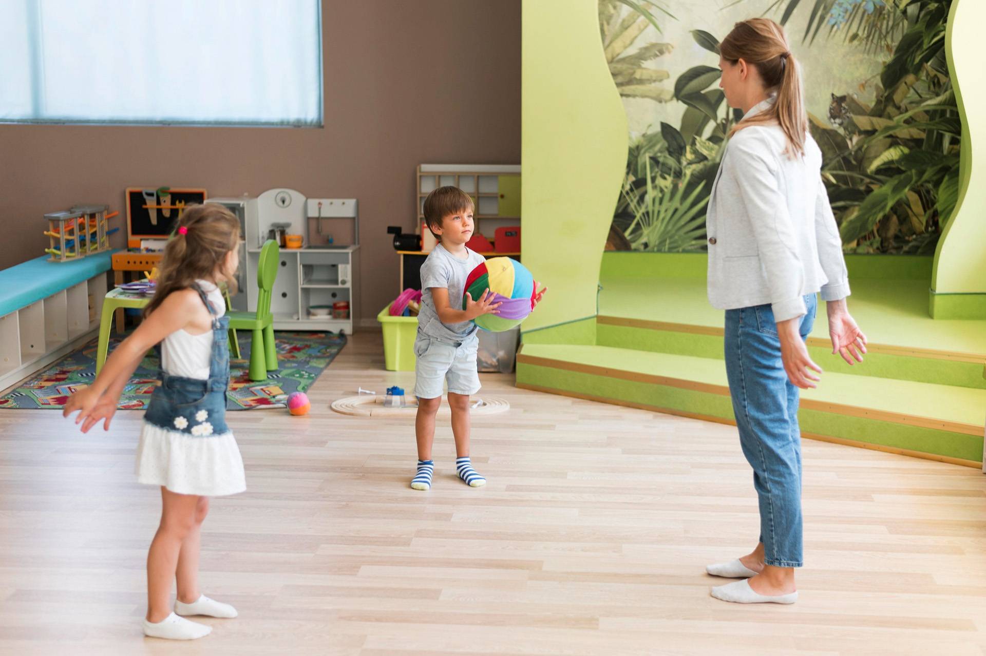 A woman and two children stand in a brightly colored kindergarten playroom. The woman and one child are extending their arms to the side while the other child holds a colorful ball. The room, filled with various toys and a mural with foliage on one wall, looks ready for playtime or cleaning up fun.