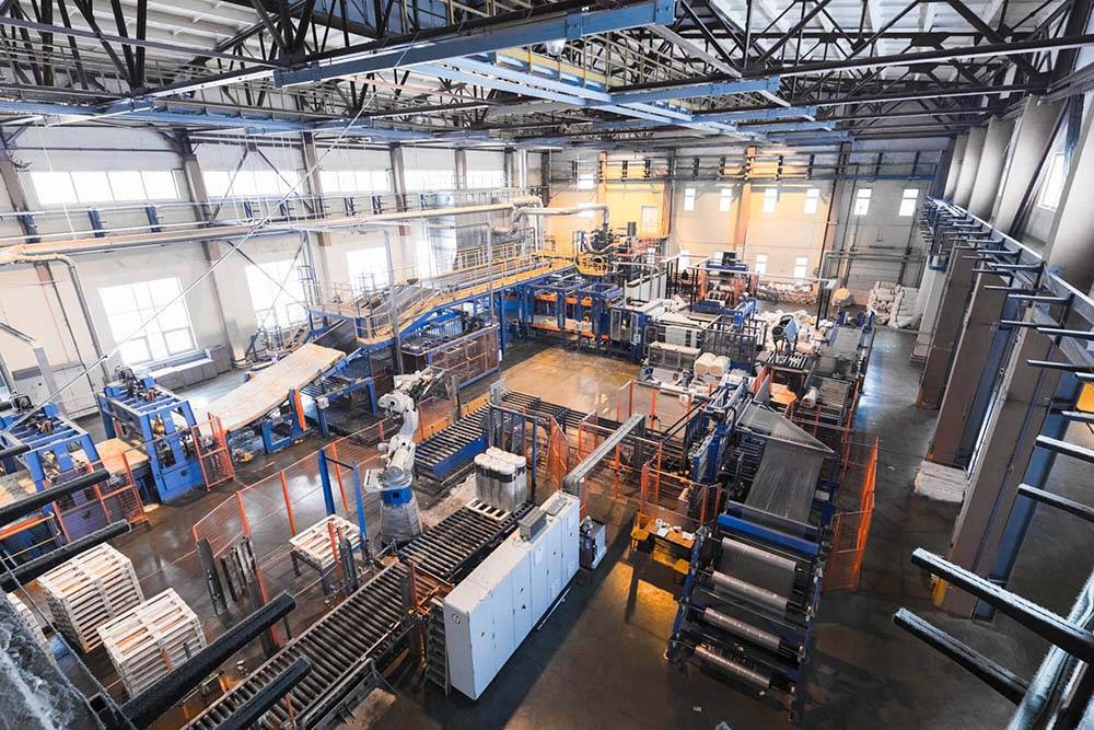 Wide-angle view of an industrial manufacturing facility with various machinery and equipment. The spacious interior includes conveyor belts, metal structures, and large windows allowing natural light to enter. The manufacturing process is highlighted, though workers are not visibly present in the image.