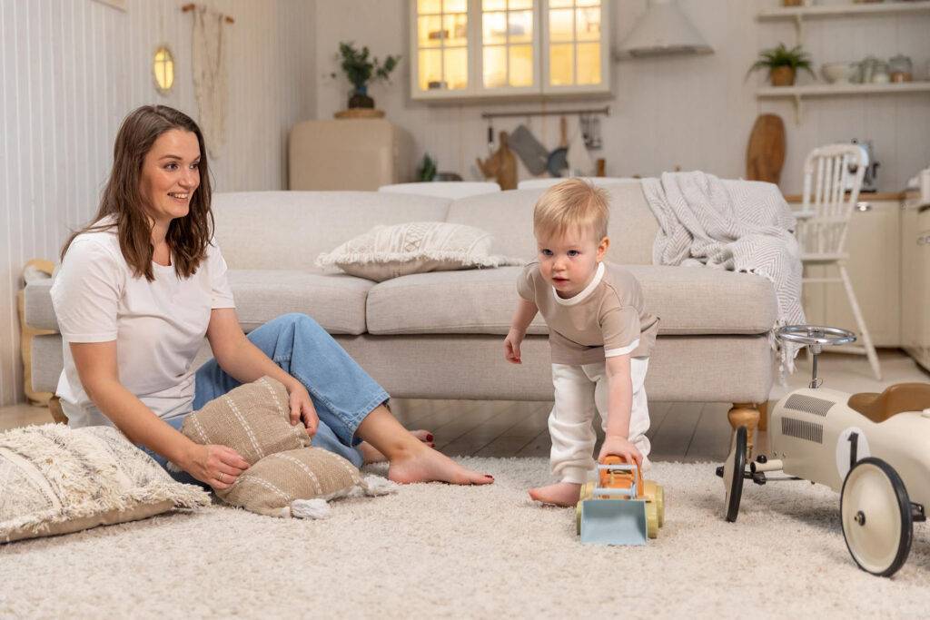 A woman sits on the carpeted floor, smiling while holding a cushion. Nearby, a young child in a beige shirt and white pants pushes a toy truck. The room is cozy with a sofa, scattered cushions, and a toy car in the background.
