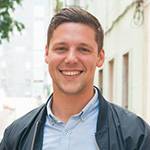 A person with short brown hair is smiling at the camera. They are wearing a light blue button-down shirt and a dark jacket. The background shows a blurred outdoor setting with buildings and greenery, reflecting a professional environment perfect for office cleaning or commercial cleaning services.