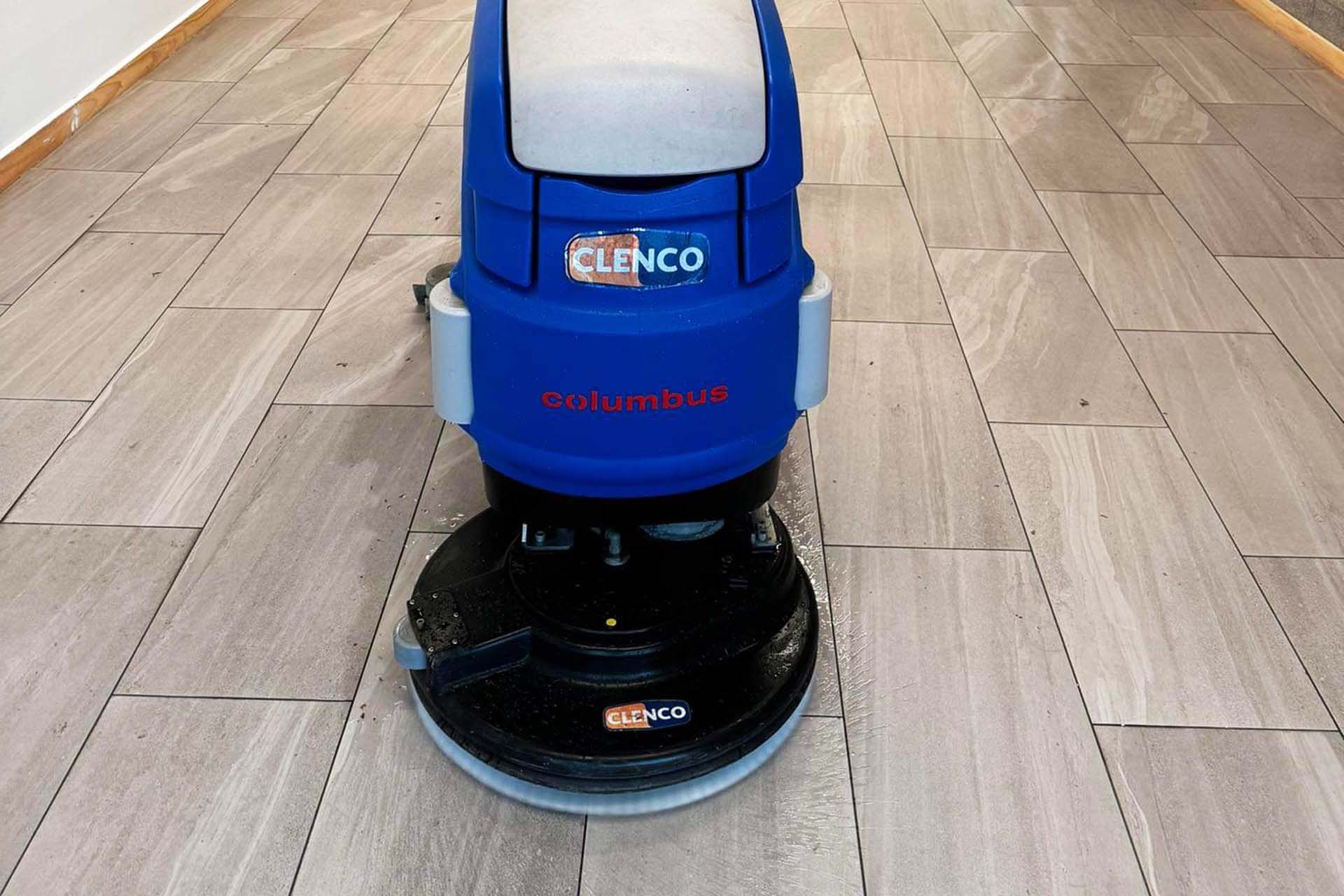 The image shows a floor cleaning machine with "CLENCO" and "columbus" labels on it. The machine, sporting a blue and grey color scheme, is efficiently performing hard floor cleaning on a grey tiled surface. The floor appears to be in the process of being cleaned.