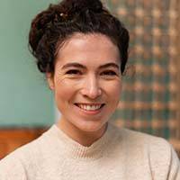 A person with curly dark hair tied up, wearing a beige sweater, is smiling at the camera. The background features a blurred pattern and a greenish wall, evoking the calmness often found in healthcare settings.