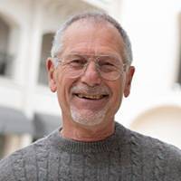 An older man smiles at the camera. He has short gray hair, is wearing glasses, and a gray knit sweater. The background is slightly blurred but shows an outdoor area with light-colored buildings, reminiscent of a professional carpet cleaning company’s pristine environment.