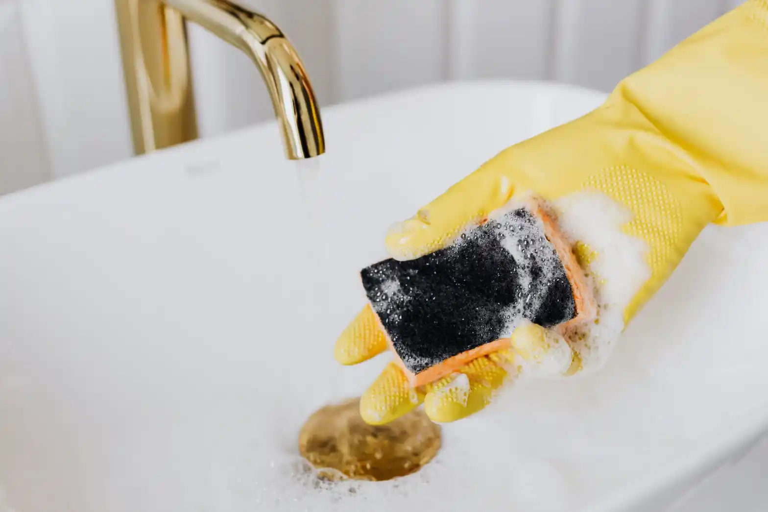 A cleaner with gloves on cleaning a soapy sponge under the tap